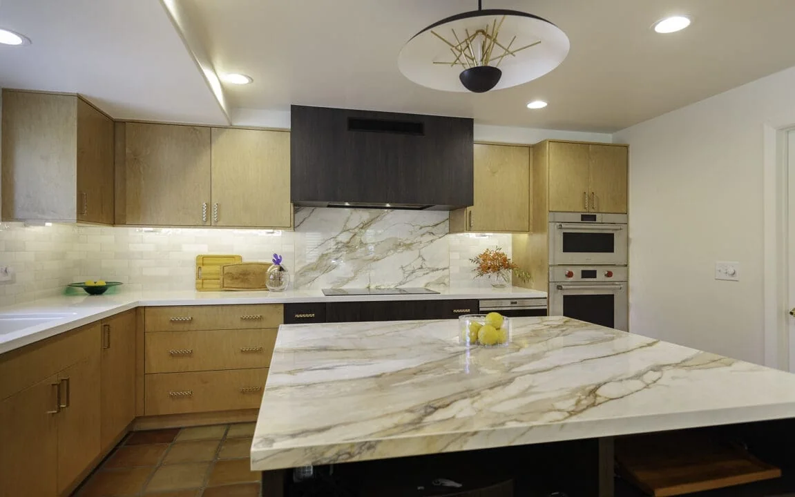Veined porcelain kitchen island countertop coordinated with the backsplash behind a dark cooktop hood and natural wood cabinets