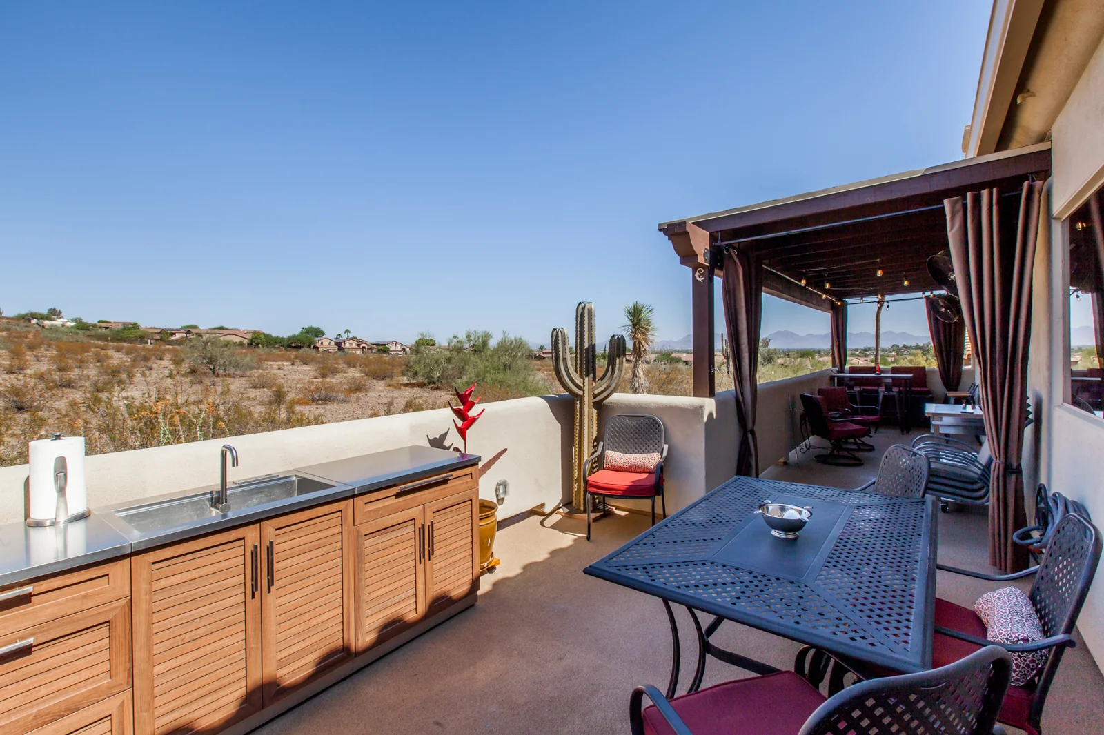 A covered outdoor patio featuring a kitchen area with a stainless steel sink, a dining table with chairs, and a desert view.