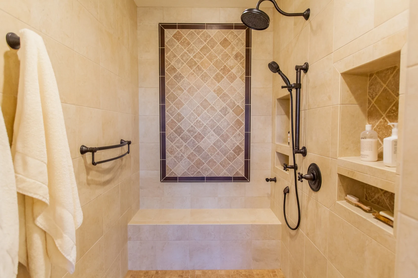 Expanded shower with porcelain wall tile, a large bench, decorative accent panel, hand shower, niches, and grab bars