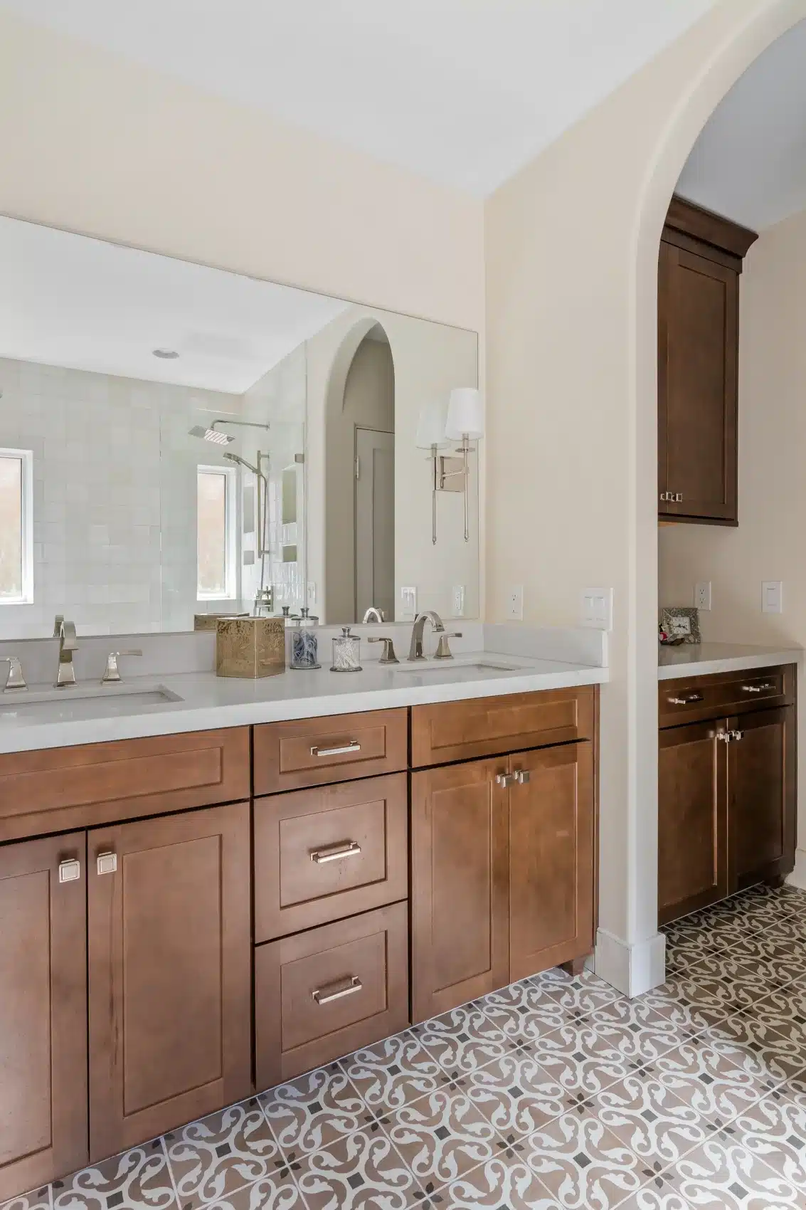 Wood double vanity beside arched opening, patterned tile floor, and light quartz countertop