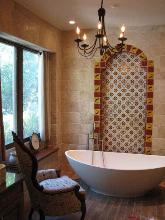 Portrait view of a freestanding white bathtub, arched decorative tile panel, dark chandelier, and large bathroom window