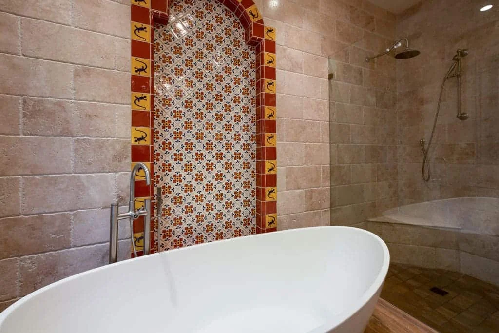 Freestanding white tub in front of an arched Mexican tile panel beside a travertine shower with a built-in bench