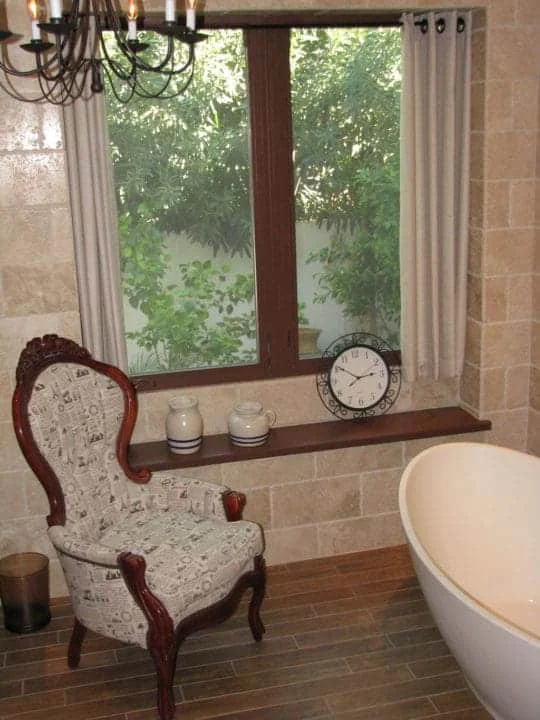 Carved wood armchair beside a large bathroom window with light curtains, travertine walls, and the edge of the freestanding tub